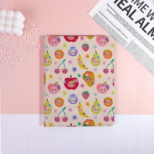 May include: A rectangular notebook with a beige cover featuring a colorful pattern of smiling fruits, including apples, pears, cherries, oranges, strawberries, and bananas. The design includes blue, purple, and yellow stars.