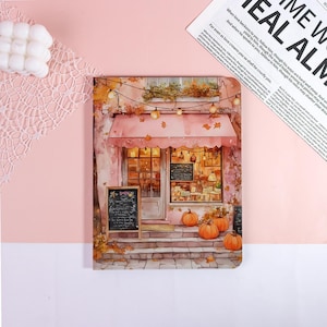 May include: A notebook with a watercolor illustration of a cafe with a pink awning, pumpkins, and string lights. The notebook has a pink background and a small chalkboard sign. The text on the page reads "EAL ALM" and "ME W".