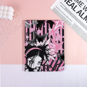 May include: A pink and black notebook featuring a cartoon illustration of a girl with headphones, stars, and hearts. The design includes black and pink stripes. The notebook is on a pink surface with a white and pink lace doily.