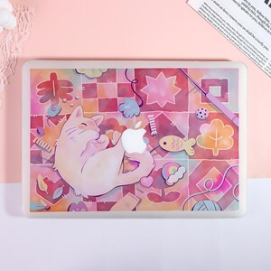 May include: A laptop skin featuring a light pink cat curled up on a colorful, grid-patterned background. The design includes various small icons like a fish, a rainbow, a ball of yarn, and stars. A white Apple logo is visible in the center.