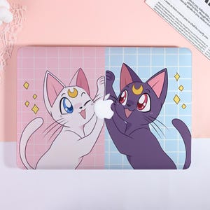 May include: Laptop case featuring a cartoon design with two cats, one white and one purple, holding an Apple logo. The background is split into pink and blue sections with a grid pattern and yellow star accents.