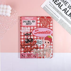 May include: A red and white checkered notebook with various patches. Patches include the phrases "Hello I am Doing My Best", "Lover Girl", "Sweetie", "Lucky Mama", and "Love of my life". There is also a strawberry baby graphic.