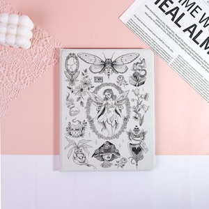 May include: A notebook with a white cover, showcasing a detailed black and white illustration. The design includes a fairy, flowers, a moth, and other mystical elements. The text on the page reads "When love beckons to you, follow him..."
