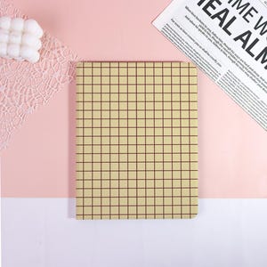 May include: A beige notebook with a grid pattern, resting on a pink surface. The grid lines are dark brown. A white, cloud-shaped object and a newspaper clipping are in the background.