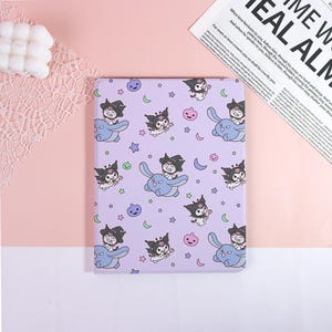 May include: A lavender notebook featuring a repeating pattern of a cartoon character with a witch hat, riding a blue creature, along with stars, moons, and jack-o'-lanterns. The notebook is rectangular and has a soft cover.