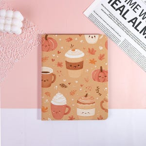 May include: A tan notebook with a repeating pattern of smiling pumpkins, coffee cups, and autumn leaves. The notebook has a soft cover and is likely used for journaling or note-taking. The background is a light pink surface.