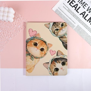 Könnte beinhalten: Eine beige Tablet-Hülle mit einem Design von drei Cartoon-Katzen, die Mützen tragen. Die Katzen sind in Orange-, Braun- und Grautönen gehalten und haben große Augen. Rosa Herzen sind um die Katzen herum verstreut. Die Hülle hat ein skurriles Design.