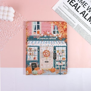 May include: A notebook with a colorful illustration of a "Pumpkin Spice" cafe. The cafe has a blue awning, a wreath on the door, and pumpkins in front. The text "Coffee Tea" is on a chalkboard. The background is pink.