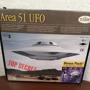May include: A Testors Area 51 UFO model kit box featuring a white saucer-shaped craft against a desert landscape. The box includes the text Area 51 UFO, Top Secret, and a Bonus Pack label for a 7 inch tall alien figure. The scale is 1:48.