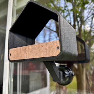 May include: A black, textured, window-mounted bird feeder with a wooden accent. The feeder has a rectangular shape with a curved top, and a small wooden panel with a wood grain pattern. It is mounted on a black bracket.