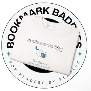 May include: A white sweatshirt featuring the text Bookmark Baddies EST. 2024, a graphic of a ghost holding blue books with a crescent moon, and the text Two Years of BOOKS, BOOS, & BADDIES. The sweatshirt is centered on a circular logo background.