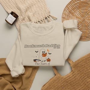 May include: A cream-colored crewneck sweatshirt featuring a graphic of a ghost reading a book next to a pumpkin, bats, and a crescent moon. The text reads "Bookmark Baddies EST. 2024" and "Two Years of" in a dark brown, retro-style font.