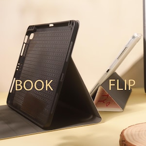 Op de afbeelding: Zwarte tablethoes met het woord "BOOK" erop gedrukt. Een witte tablet staat op een driehoekige standaard met het woord "FLIP" erop gedrukt. De hoes is open en onthult een gestructureerd interieur.