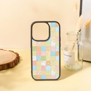 May include: A phone case with a patchwork design in pastel colors. The case has a black border and a cut-out for the camera. The patchwork pattern includes squares of various colors and patterns, such as polka dots and floral prints.