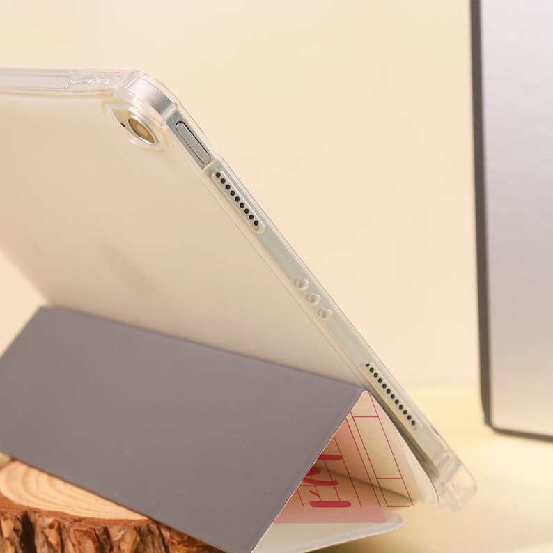 Op de afbeelding: Een doorzichtige beschermhoes voor een tablet, met een grijze hoes die is opgevouwen om het apparaat te ondersteunen. De tablet is wit met een zwarte rand. De hoes is transparant.
