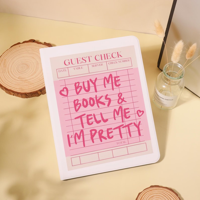 Op de afbeelding: Een wit notitieblok met een roze gastenbonontwerp. Het notitieblok heeft de tekst "BUY ME BOOKS & TELL ME I'M PRETTY" in het roze. Het notitieblok ligt op een lichtgele ondergrond.