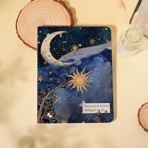 May include: A celestial-themed journal featuring a crescent moon, sun, and stars against a dark blue sky. The cover includes the text "I am out with lanterns looking for myself". The journal is rectangular and has a decorative design.