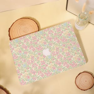 Pu&ograve; includere: Una custodia per laptop con un motivo floreale rosa, giallo e blu su sfondo verde chiaro. La custodia presenta un logo Apple bianco. La custodia per laptop &egrave; su una superficie giallo chiaro con elementi in legno.