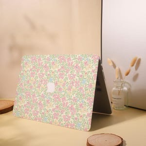 Pu&ograve; includere: Un laptop con una custodia a fantasia floreale. La custodia presenta un motivo di fiori rosa, gialli e blu su uno sfondo giallo chiaro. Il logo Apple &egrave; visibile.