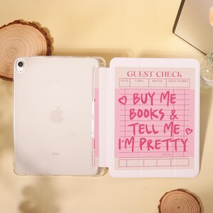Op de afbeelding: Een doorzichtige iPad in een beschermhoes naast een roze notitieblok. Het notitieblok leest "GUEST CHECK" bovenaan, met de tekst "BUY ME BOOKS & TELL ME I'M PRETTY" in roze. De iPad heeft het Apple-logo.
