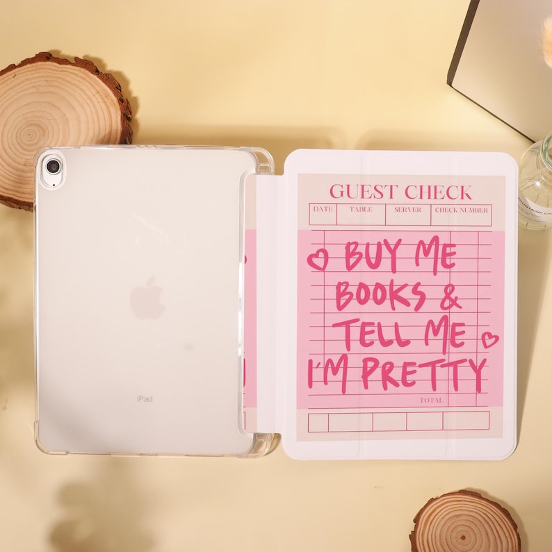 Op de afbeelding: Een doorzichtige iPad in een beschermhoes naast een roze notitieblok. Het notitieblok leest "GUEST CHECK" bovenaan, met de tekst "BUY ME BOOKS & TELL ME I'M PRETTY" in roze. De iPad heeft het Apple-logo.