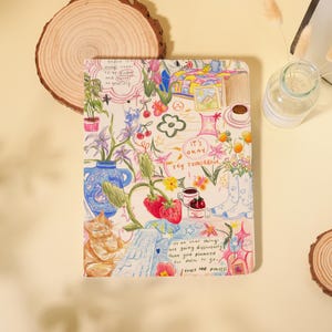 May include: A colorful notebook with a whimsical illustration. The cover features drawings of flowers, strawberries, a cat, and text that reads "It's okay, try tomorrow." The notebook is surrounded by wooden slices and a glass jar.