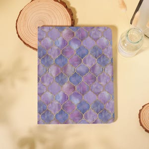 May include: A notebook with a watercolor-style pattern in shades of purple and blue, accented with gold-colored outlines. The design features a repeating quatrefoil motif. The notebook is rectangular and appears to be closed.