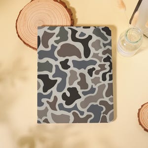 May include: A rectangular notebook featuring a camouflage pattern in shades of dark gray, light gray, and brown. The cover has a smooth finish with rounded corners, displaying an abstract organic shape design across the entire front surface.