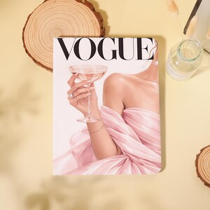 May include: A rectangular wall art print featuring the word VOGUE in bold black letters at the top. The illustration depicts a person in a flowing light pink gown holding a stemmed cocktail glass, wearing a ring and a delicate gold bracelet.