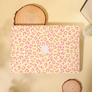 May include: A rectangular laptop cover featuring a repeating pink leopard print pattern on a pale yellow background. The cover has a centered cutout revealing the white Apple logo. The design covers the entire top surface of the laptop shell.