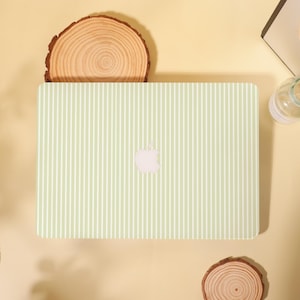 May include: A light green laptop featuring a white vertical stripe design. The Apple logo is centrally placed. The laptop rests on a pale yellow surface, with wooden slices as decoration.