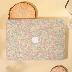 Pu&ograve; includere: Una custodia per laptop con un motivo floreale in rosa, giallo e blu su sfondo verde chiaro. La custodia presenta un logo Apple bianco. Si trova su una superficie gialla, accompagnata da una fetta di legno e altri oggetti.