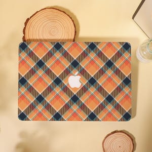 May include: A laptop with a colorful plaid pattern in orange, brown, navy, and teal. The Apple logo is visible in white. The laptop is on a light yellow surface, with a wooden slice and other objects in the background.