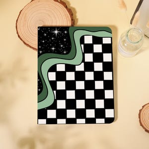 May include: A rectangular notebook with a black and white checkered pattern on the right side. The left side features a black starry sky design with a green wavy border. The notebook is on a light yellow surface.