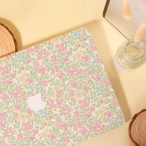 Pu&ograve; includere: Una custodia per laptop con un motivo floreale nei toni pastello di fiori rosa, gialli e blu su uno sfondo verde chiaro. La custodia ha un logo Apple bianco. La custodia &egrave; progettata per proteggere un laptop.