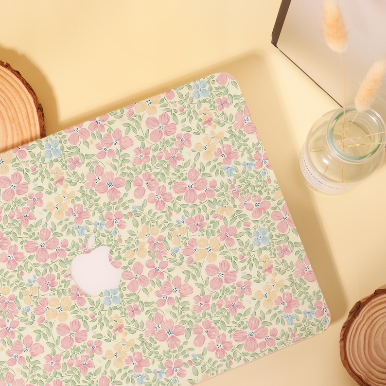 Pu&ograve; includere: Una custodia per laptop con un motivo floreale nei toni pastello di fiori rosa, gialli e blu su uno sfondo verde chiaro. La custodia ha un logo Apple bianco. La custodia &egrave; progettata per proteggere un laptop.