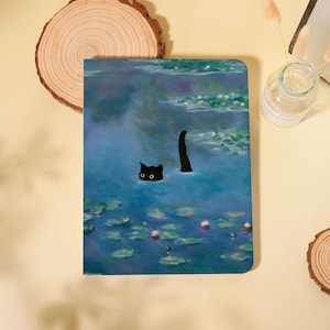 May include: A notebook featuring a whimsical illustration of a black cat in a pond, with only its head and tail visible above the water. The design is reminiscent of a Monet painting, with a blue and green color palette.