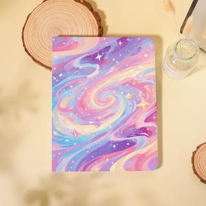 May include: A rectangular notebook with a colorful, swirling galaxy design in shades of purple, pink, blue, and yellow. The cover features stars and a dreamy, artistic aesthetic. The notebook is set on a light yellow surface.