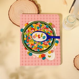 May include: A rectangular notebook with a pink grid pattern cover featuring a graphic illustration of a green bowl filled with colorful ring-shaped cereal in milk. A blue spoon rests in the bowl with the word HELLO printed on the spoon bowl.