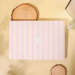 May include: A laptop with a pastel striped design in shades of pink, peach, and cream. The Apple logo is visible on the cover. The laptop is placed on a light yellow surface with wooden accents.