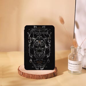 May include: Black tablet case featuring a silver skull design with the words "Morsmordre" and "Death Eater". The case is displayed on a wooden stand, with a clear glass vase and a decorative plant in the background.