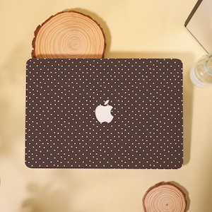 Peut inclure: Une coque d'ordinateur portable marron avec un motif à pois blancs et un logo Apple blanc. La coque est rectangulaire et recouvre le dessus de l'ordinateur portable. Le fond est de couleur jaune clair.