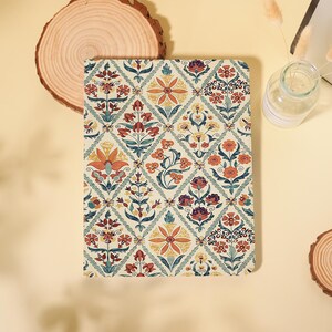 May include: A rectangular notebook featuring a cream-coloured cover with a repeating diamond pattern. Each diamond contains intricate floral illustrations in shades of orange, yellow, teal, and dark red. The design is symmetrical and botanical.