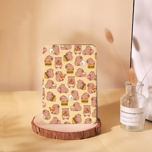 May include: A rectangular case featuring a repeating pattern of cartoon hamsters with various foods like burgers, pizza, and ice cream on a light yellow background. The case is displayed on a wooden stand.