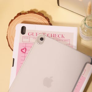 Op de afbeelding: Een doorzichtige iPad-hoes over een roze en wit gastencontrole-notitieblok. Op het notitieblok staan de woorden "GUEST CHECK" gedrukt, samen met lijnen om op te schrijven. Een houten schijf staat op de achtergrond.
