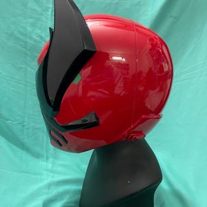 May include: A red and black plastic helmet with a pointed crest. The helmet is on a black display stand.