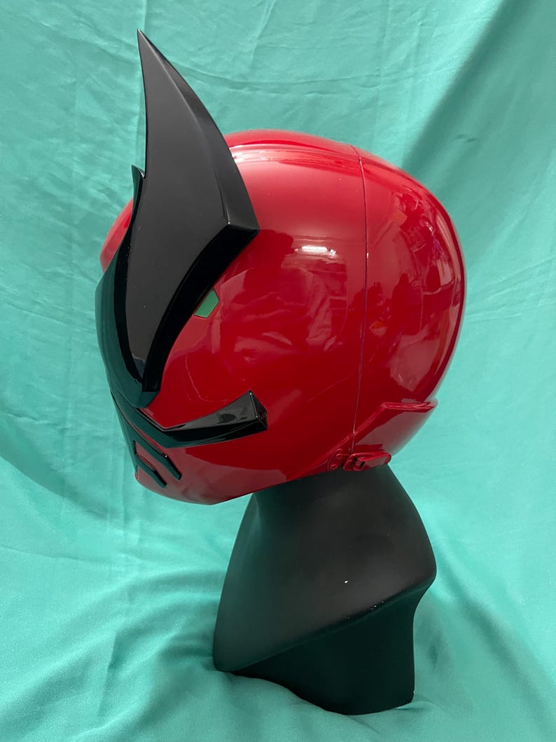 May include: A red and black plastic helmet with a pointed crest. The helmet is on a black display stand.