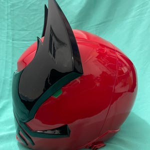 May include: A red and black plastic helmet with a pointed black crest. The helmet has a green visor and a black design on the front.