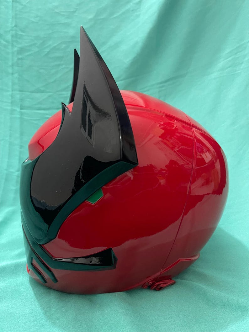 May include: A red and black plastic helmet with a pointed black crest. The helmet has a green visor and a black design on the front.