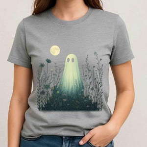 Puede incluir: Una camiseta gris con una ilustración caprichosa de un fantasma en un campo de flores bajo la luna llena. El fantasma está representado con una sábana blanca fluida y dos ojos ovalados. Las flores son en tonos de azul, amarillo y verde.